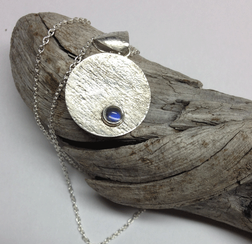 A reticulated silver disk complimented with a moonstone. This photo was taken show off the blue schiller of this lovely little moonstone. The bail also has reticulated texture. Disk is 20mm. All silver is sterling. Comes with an 18" sterling silver chain. $47